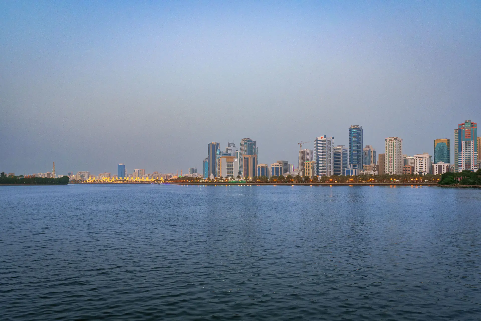 The Sharjah skyline reflected on calm water at dusk.