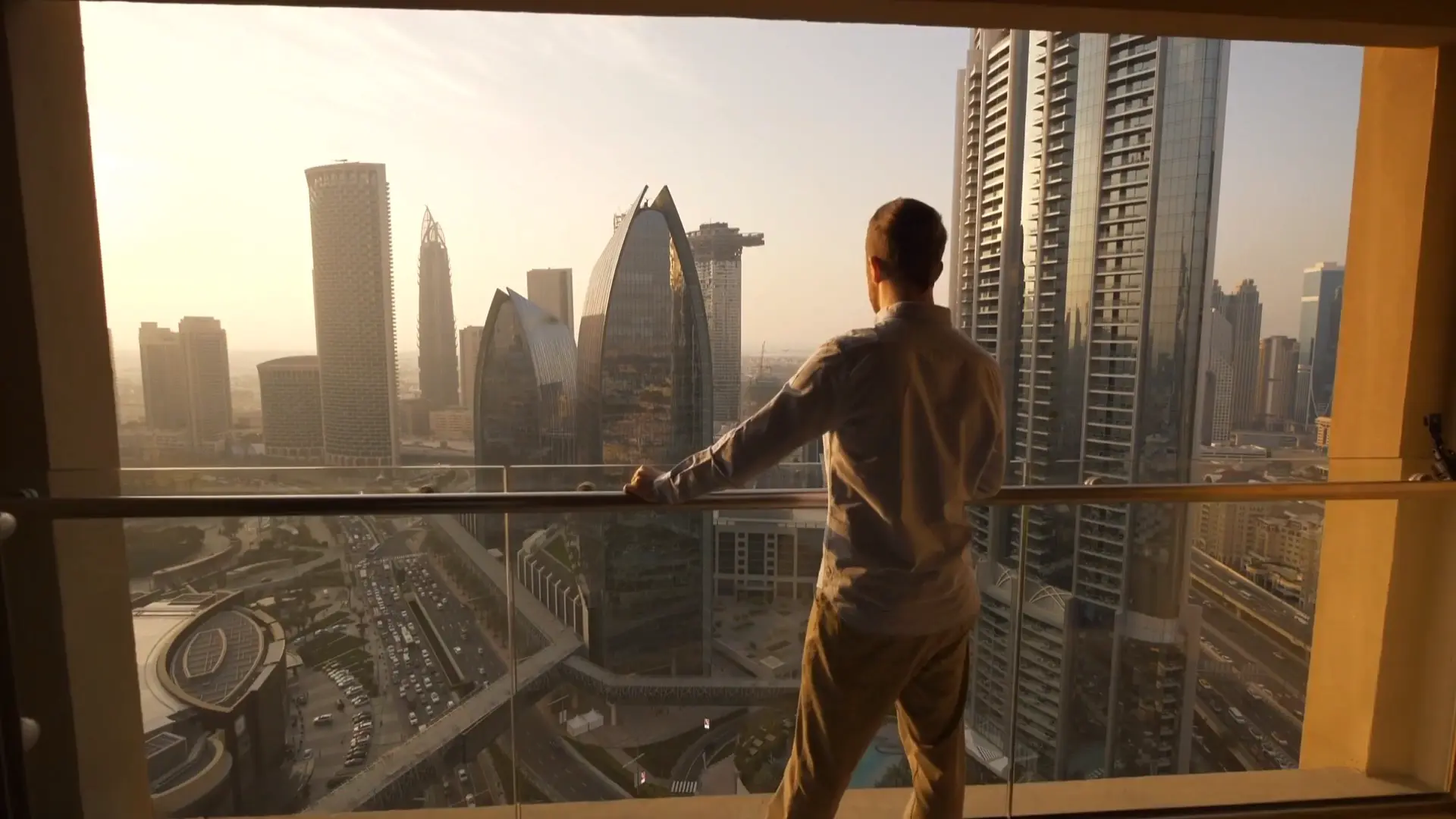Golden hour over Downtown Dubai from a high balcony, a man at the rail looking out across Sheikh Zayed Road.